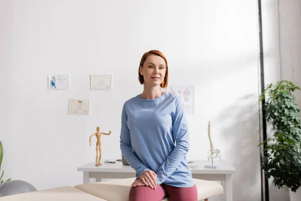 Mujer feliz sentada en la mesa de masaje en el centro de rehabilitación y sonriendo a la cámara - foto de stock