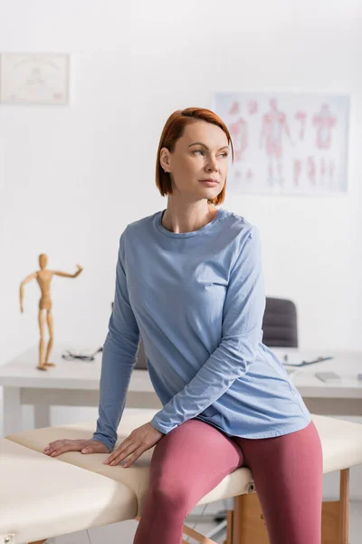 Redhead woman looking away while sitting on massage table in consulting room of recovery center — Stock Photo