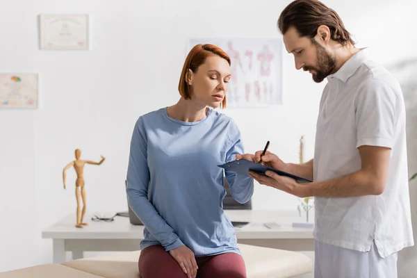 Redhead woman near bearded physiotherapist writing diagnosis on clipboard in consulting room — Stock Photo