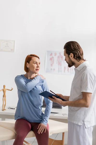Woman touching painful shoulder during consultation with physiotherapist holding clipboard in consulting room — Stock Photo