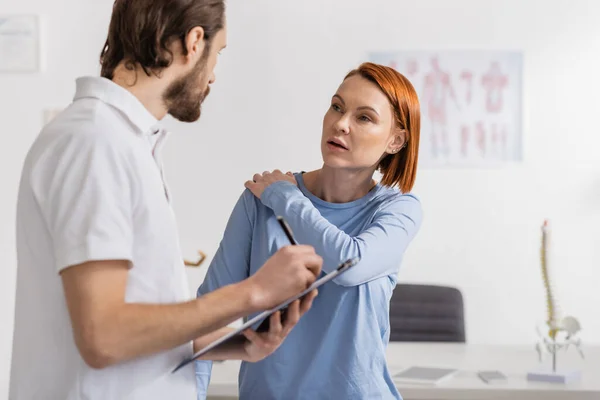 Руда жінка з травмованим плечем розмовляє з фізіотерапевтом, пишучи діагноз на кишені в лікарні — стокове фото