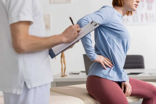 Cropped view of physiotherapist with clipboard writing diagnosis near woman suffering from pain in loin in consulting room — Stock Photo