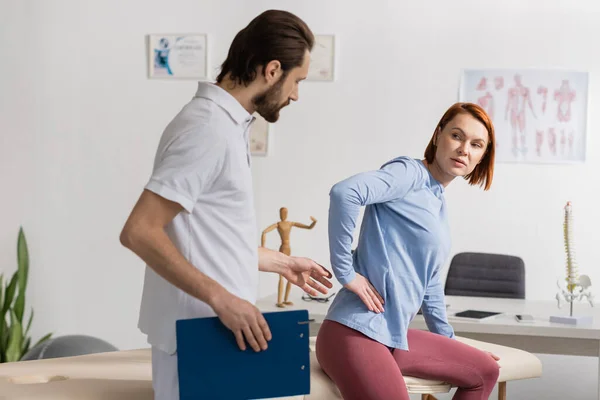 Bearded physiotherapist with clipboard near woman touching painful loin in consulting room — Stock Photo