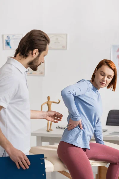 Redhead woman touching painful loin near manual therapist with clipboard in consulting room — Stock Photo