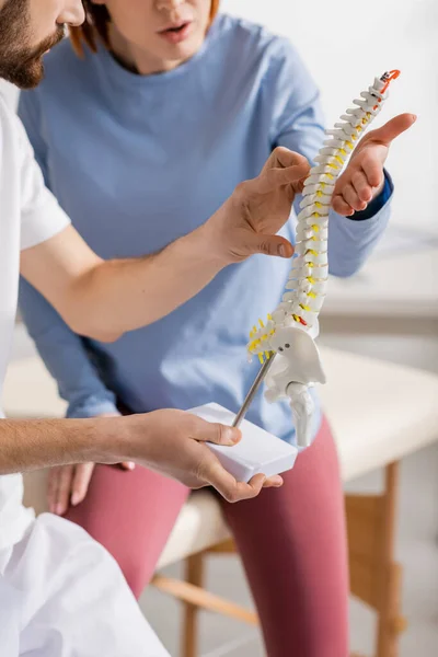 Vista recortada del quiropráctico apuntando al modelo de columna vertebral cerca del paciente en el centro de rehabilitación - foto de stock