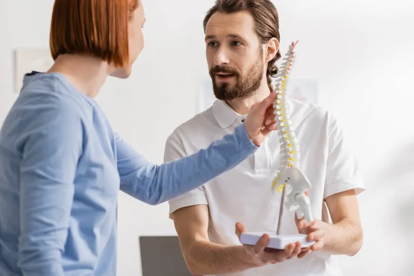 Ostéopathe barbu tenant modèle de colonne vertébrale et parlant à la rousse femme dans la salle de consultation — Photo de stock