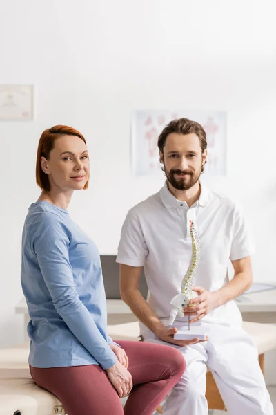 Felice rossa donna e fisioterapista sorridente con modello di colonna vertebrale guardando la fotocamera in sala di consultazione — Foto stock