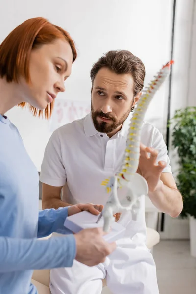 Redhead woman holding spine model near bearded chiropractor in consulting room — Stock Photo