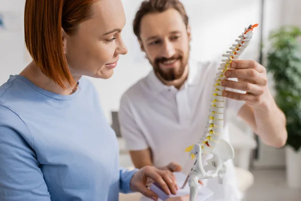Chiropraticien souriant montrant le modèle de colonne vertébrale à la femme rousse lors d'un rendez-vous dans la salle de consultation — Photo de stock