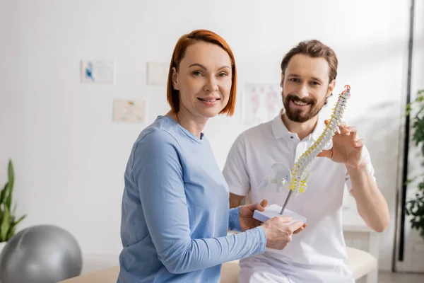 Cheerful redhead woman and bearded chiropractor looking at camera near spine model in consulting room — Stock Photo