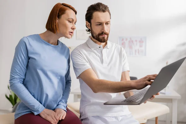 Barbuto terapeuta manuale mostrando laptop alla donna rossa durante la consultazione nel centro di riabilitazione — Foto stock