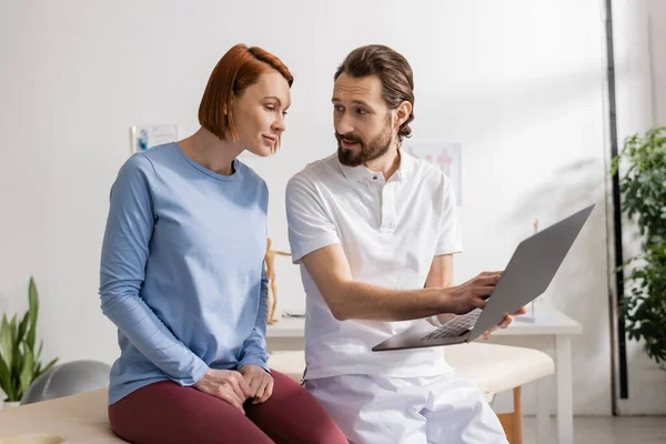 Fisioterapeuta barbudo com laptop conversando com mulher sorridente durante consulta no centro de reabilitação — Fotografia de Stock