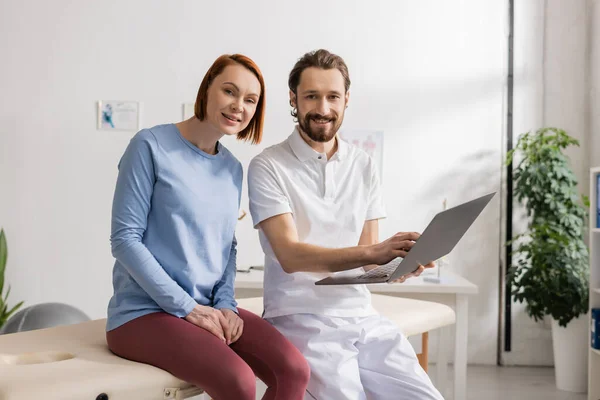 Mujer positiva y fisioterapeuta barbuda con portátil mirando a la cámara mientras está sentado en la mesa de masaje en la sala de consulta - foto de stock