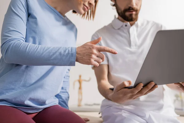 Partial view of woman pointing at laptop near bearded physiotherapist in consulting room — Stock Photo
