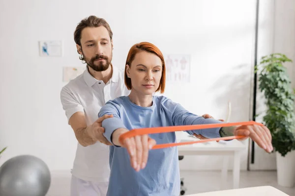 Bearded chiropractor assisting redhead woman training arms with resistance band in clinic — Stock Photo