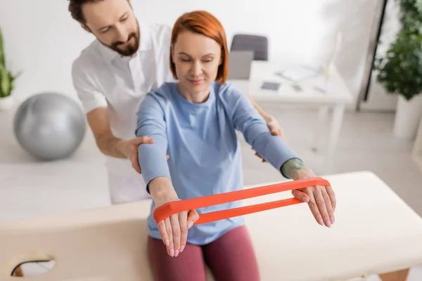 Physiotherapist supporting smiling woman working out with resistance band on massage table in clinic — Stock Photo