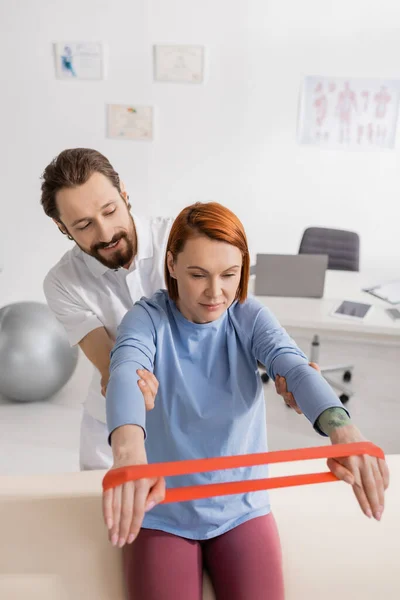 Smiling physiotherapist supporting redhead woman training arms with resistance band in rehab center — Stock Photo