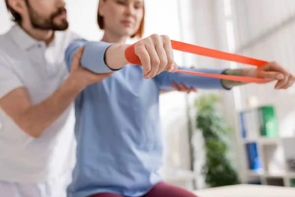 Cropped view of blurred rehabilitation trainer supporting woman exercising with elastics in consulting room — Stock Photo