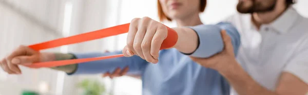 Cropped view of physiotherapist supporting arms of woman training with resistance band in rehab center, banner — Stock Photo
