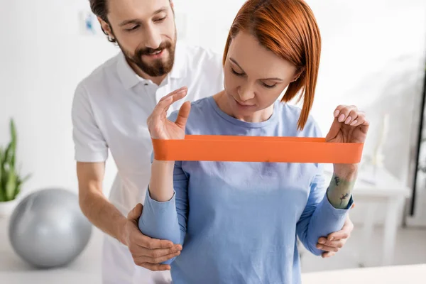 Redhead woman training arms with resistance band near bearded physiotherapist in rehab center — Stock Photo