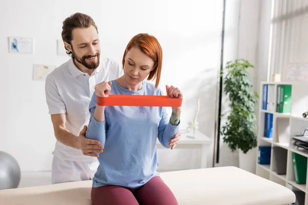 Redhead woman sitting on massage table and exercising with resistance band near positive physiotherapist in hospital — Stock Photo