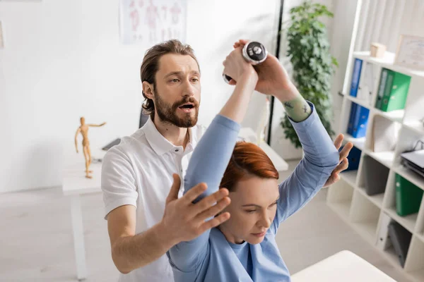 Rehabilitation doctor helping redhead woman exercising with dumbbell in recovery center — Stock Photo