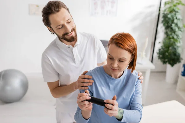 Positive Physiotherapeutin umarmt Schultern einer rothaarigen Frau, die im Sprechzimmer auf ihr Handy schaut — Stockfoto