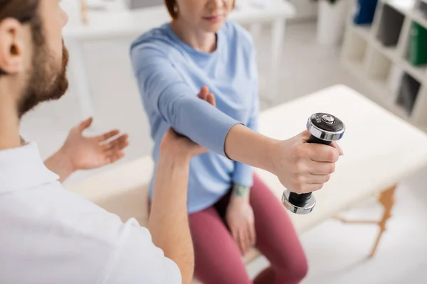 Visão parcial do braço de treinamento da mulher borrada com haltere perto de especialista em reabilitação na clínica — Fotografia de Stock