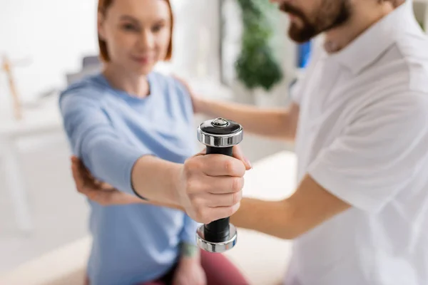 Foco seletivo do haltere na mão da mulher que exercita perto do fisioterapeuta borrado no centro de reabilitação — Fotografia de Stock