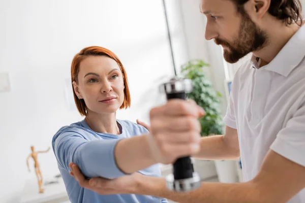 Bearded physiotherapist helping redhead woman working out with dumbbell in rehab center — Stock Photo