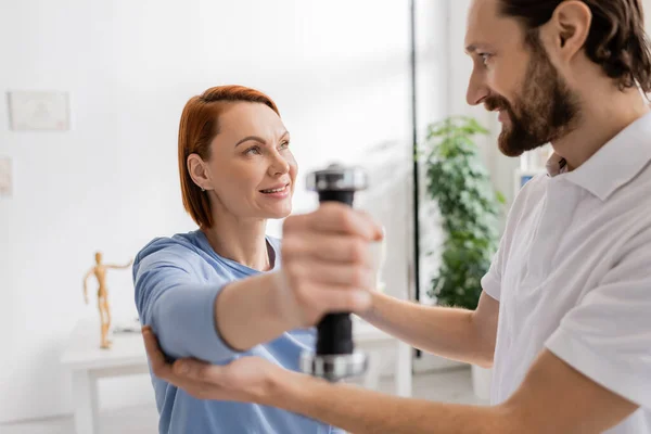 Happy woman looking at physiotherapist while training arm with dumbbell in rehab center — Stock Photo