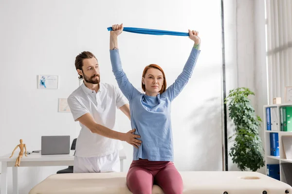 Bearded rehabilitation doctor assisting woman sitting on massage table in consulting room and training arms with resistance band — Stock Photo
