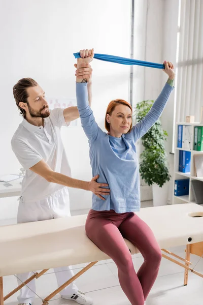Redhead woman sitting on massage table and training arms with resistance band near bearded physiotherapist in clinic — Stock Photo