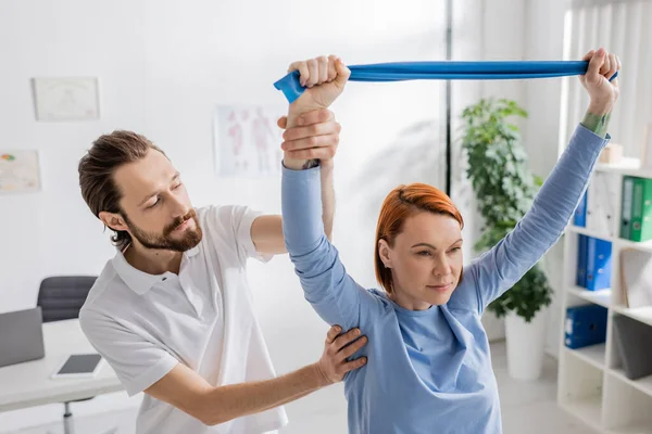 Rehabilitologist assisting redhead woman with resistance band training arms in consulting room — Stock Photo