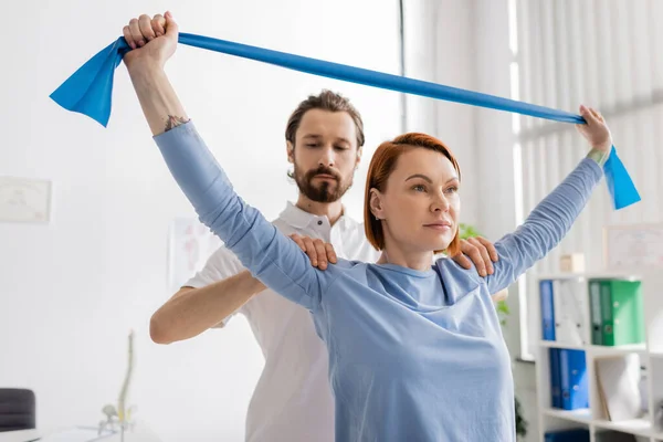 Bärtiger Physiotherapeut hilft Patientin beim Training mit Widerstandsband im Reha-Zentrum — Stockfoto