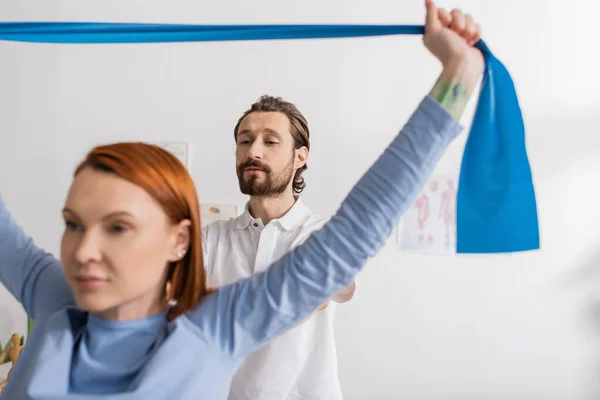 Bearded physiotherapist looking at redhead woman exercising with resistance band on blurred foreground in recovery center — Stock Photo