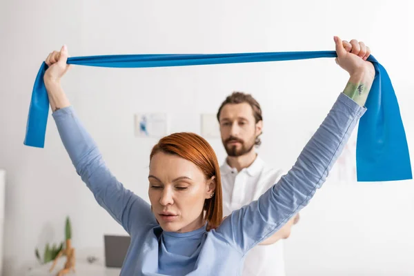 Redhead woman working out with elastics near blurred rehabilitologist in recovery center — Stock Photo