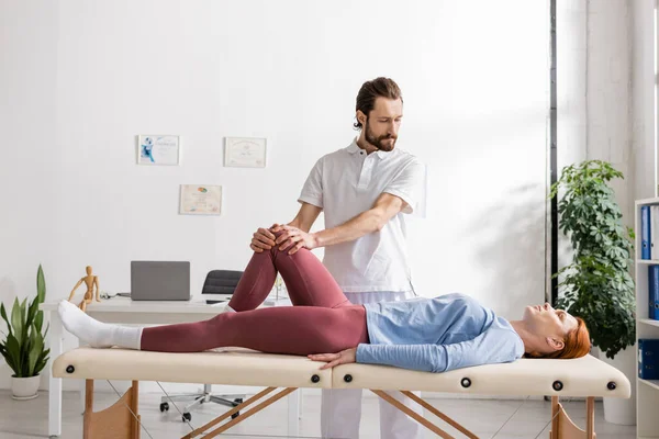 Barbuto terapeuta manuale facendo massaggio al ginocchio alla donna nel centro di riabilitazione — Foto stock