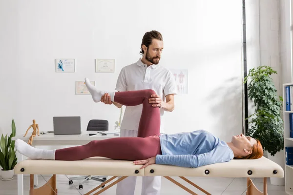 Reabilitologista flexionando a perna dolorosa da mulher deitada na mesa de massagem no consultório — Fotografia de Stock