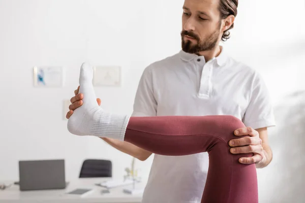 Bearded osteopath flexing injured leg of patient during rehabilitation in clinic — Stock Photo