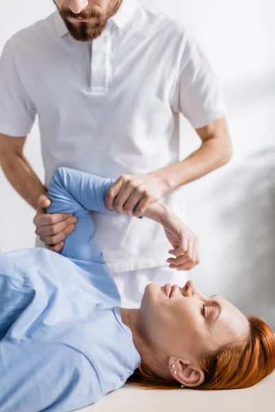 Fisioterapeuta barbudo flexionando braço doloroso da mulher durante terapia de reabilitação na clínica — Fotografia de Stock