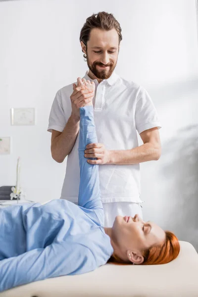 Fisioterapeuta sonriente estirando el brazo de la mujer mientras hace masajes para aliviar el dolor en el centro de recuperación - foto de stock