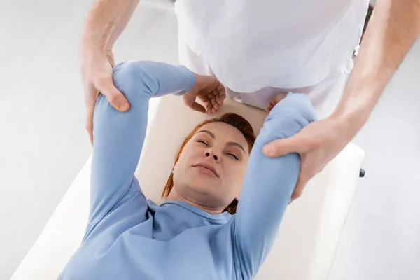Visão de alto ângulo da mulher com olhos fechados perto do fisioterapeuta massagear braços dolorosos na clínica — Fotografia de Stock