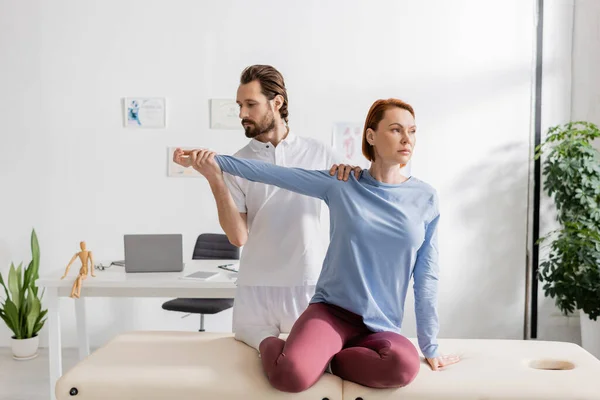 Rehabilitologist examining arm of redhead woman sitting on massage table in consulting room — Stock Photo