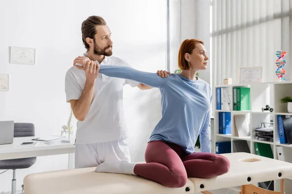 Chiropractor stretching arm of redhead woman sitting on massage table in rehabilitation center — Stock Photo