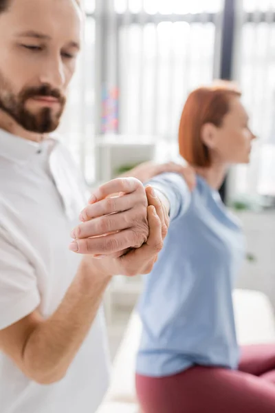 Bearded physiotherapist stretching painful arm of blurred woman during diagnostics in consulting room — Stock Photo