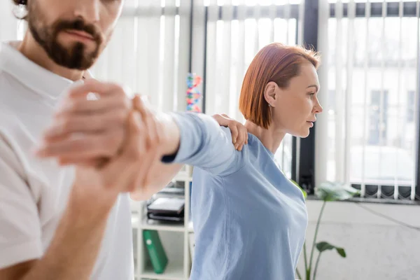 Redhead woman with injured arm near blurred physiotherapist doing diagnostics in consulting room — Stock Photo