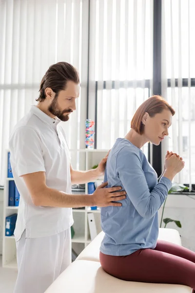 Visão lateral do fisioterapeuta barbudo tocando as costas dolorosas da mulher ruiva durante a consulta no consultório — Fotografia de Stock