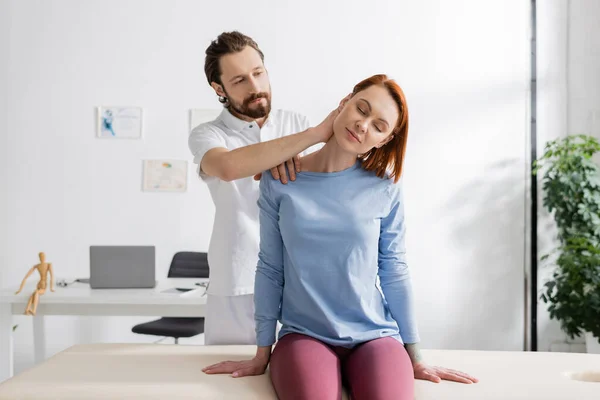 Бородатий фізіотерапевт вивчає болючу шию жінки, що сидить на масажному столі в консалтинговій кімнаті — стокове фото