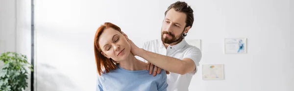 Bearded chiropractor examining injured neck of redhead woman in consulting room, banner — Stock Photo